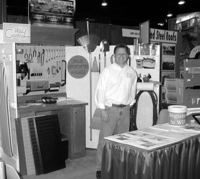 Black and white photo of a man standing in a trade show booth with various products and signs.