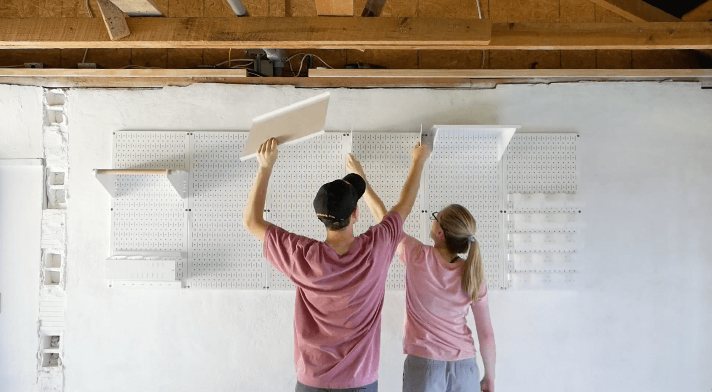 Two people installing a pegboard in a room with wooden beams.